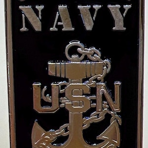 May include: A rectangular black metal emblem with the words "UNITED STATES NAVY" in silver lettering. Below, a silver anchor design with the letters "USN" is visible. The emblem has a gold border.