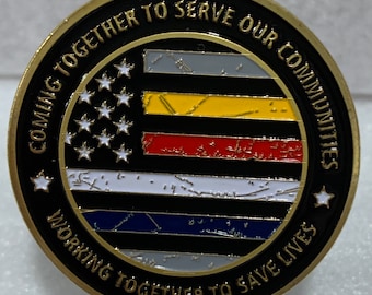 Puede incluir: Una moneda de desafío de color dorado con un fondo negro y un borde fino de oro. El centro presenta una delgada bandera estadounidense en blanco y negro con una franja roja, amarilla y azul en el medio. El texto "Coming Together to Serve Our Communities Working Together to Save Lives" está escrito en un círculo dorado alrededor de la bandera.