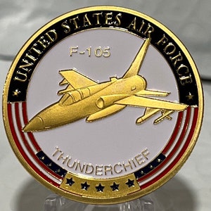 May include: A gold-colored coin with a white center featuring a gold F-105 Thunderchief jet plane. The coin is surrounded by a red, white, and blue American flag design with the words "United States Air Force" in black lettering.