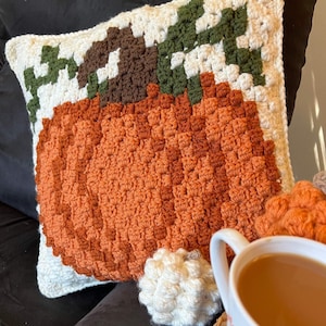 Op de afbeelding: Een vierkant gehaakt kussen met een groot oranje pompoenontwerp met een bruine steel en groene bladeren op een crèmekleurige achtergrond. De textuur is gemaakt met dichte, verhoogde steken. Een kleine witte gehaakte bol ligt ernaast.
