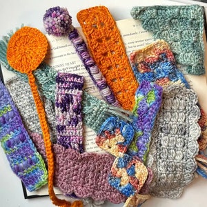 May include: A collection of handmade crocheted bookmarks in various colors and patterns, including purple, orange, gray, pink, and blue. The bookmarks are arranged on a book with the text "WILD" and "MININE HEART" visible.