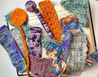 May include: A collection of various crocheted bookmarks in assorted colors and textures, including solid orange, variegated purple, blue, and pink yarns, featuring different stitch patterns, tassels, and a pom-pom, all resting on an open book page.