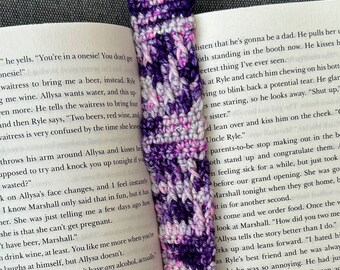 May include: A rectangular crochet bookmark featuring a variegated yarn pattern in shades of deep purple, light lavender, and white. The bookmark is placed vertically between the pages of an open book with visible black printed text.