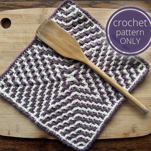 Könnte beinhalten: Ein lila-weißes Häkel-Geschirrtuch mit einem Zickzackmuster. Das Geschirrtuch liegt auf einem Holz-Schneidebrett mit einem Holzlöffel darauf.