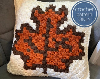 May include: A square crochet cushion cover featuring a large maple leaf design in dark brown and burnt orange against a cream-coloured background. A blue circular graphic in the top right corner contains the white text crochet pattern ONLY.