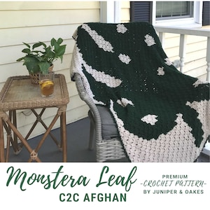 Puede incluir: Una manta de crochet verde y blanca con un patrón de hoja de monstera. La manta está drapeada sobre una silla de mimbre en un porche. El texto "Monstera Leaf C2C Afghan - Premium Crochet Pattern - By Juniper & Oakes" está en la parte inferior de la imagen.
