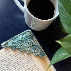 Può includere: Un segnalibro all'uncinetto in tonalità di blu e verde, con un bordo a festoni, è posizionato su un libro aperto. Una tazza di caffè bianca con caffè nero è visibile sullo sfondo.