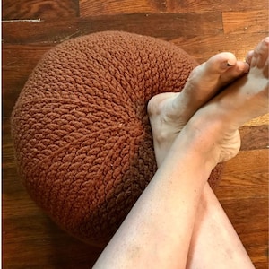 May include: A brown knitted pouf with a woven texture. A person's legs are resting on top of the pouf.