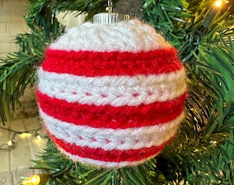 May include: A spherical Christmas tree ornament covered in a crocheted yarn cozy featuring alternating horizontal stripes of bright red and white. The ornament has a metallic silver cap at the top with a small wire loop for hanging on a tree branch.