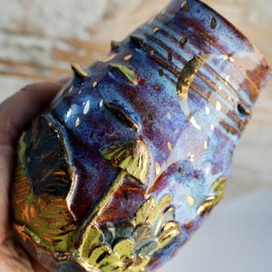 May include: A small, hand-painted ceramic vase with a blue, purple, and gold glaze. The vase has a textured surface with raised bumps and a design of mushrooms and leaves.