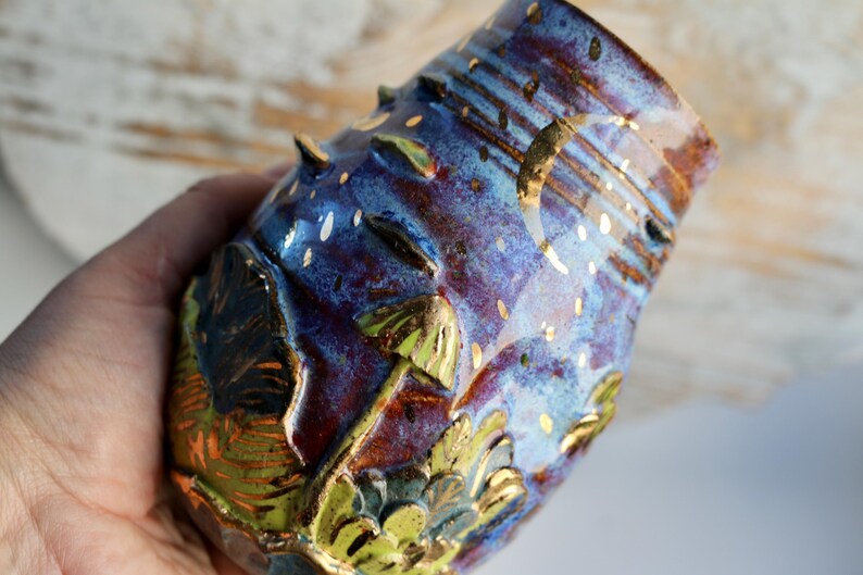 May include: A small, hand-painted ceramic vase with a blue, purple, and gold glaze. The vase has a textured surface with raised bumps and a design of mushrooms and leaves.