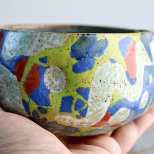 May include: A hand-held ceramic bowl with a colorful, abstract design. The bowl features a speckled, textured surface with a mix of green, blue, red, and white shapes. The rim is a light brown color.