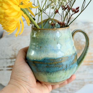 May include: A ceramic wall pocket vase with a handle, featuring a glossy glaze in shades of light green, teal, and blue. The vase has a rounded body and a small hole at the top for hanging, and it is filled with a bright yellow flower and dried stems.