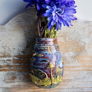 May include: A blue and purple ceramic wall vase with a mushroom and leaf design. The vase is mounted on a light wood surface and has a bouquet of purple flowers in it.