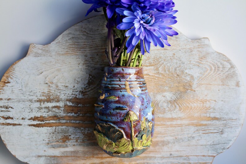 May include: A blue and purple ceramic wall vase with a mushroom and leaf design. The vase is mounted on a light wood surface and has a bouquet of purple flowers in it.
