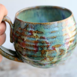 Könnte beinhalten: Eine handgefertigte Keramiktasse mit runder Form und großem Henkel. Die Tasse hat eine strukturierte Oberfläche mit einer Mischung aus Blau, Grün und Braun. Der Innenraum ist ein leuchtendes Blaugrün. Ein einzigartiges Stück.