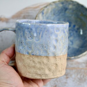 May include: Handmade ceramic mug with a textured, light blue upper half and a tan lower half. The mug has a curved handle and is paired with a matching blue bowl. The pottery has a rustic, artisanal feel.