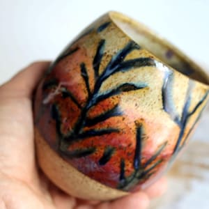 May include: Handmade ceramic mug with a unique design. The mug features a beige base and a colorful pattern of orange, red, and yellow with black branch-like shapes. The interior is a light beige color.