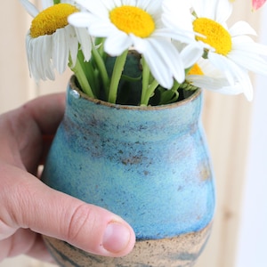 May include: A small ceramic vase with a rounded body, featuring a vibrant blue glazed upper section and a speckled beige lower base with dark horizontal markings, filled with a bouquet of white daisies and a single orange lily flower.