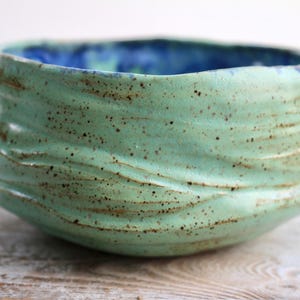May include: A handmade ceramic bowl with a textured, wavy exterior in a seafoam green color. The interior is a deep blue. The bowl has a rustic, organic aesthetic, with visible speckles and a unique shape, perfect for serving or display.