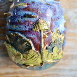 May include: A ceramic egg-shaped ornament with a blue, purple, and red glaze. The ornament features a gold-painted mushroom with a green cap and a leafy design.