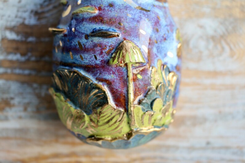 May include: A ceramic egg-shaped ornament with a blue, purple, and red glaze. The ornament features a gold-painted mushroom with a green cap and a leafy design.
