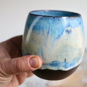 Puede incluir: Una taza de cerámica con un esmalte azul y blanco. La taza tiene una forma redondeada y un borde marrón oscuro.