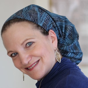 May include: A woman wearing a blue and white patterned head covering with a gold leaf-shaped earring. She is smiling and looking at the camera.