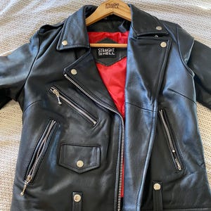 May include: Black leather jacket with a red interior lining. The jacket features a classic asymmetrical zipper closure, multiple zippered pockets, and silver hardware. The text "STRAIGHT TO HELL" is visible on the hanger.