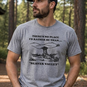 Puede incluir: Una camiseta gris jaspeada con el texto "THERE'S NO PLACE I'D RATHER BE THAN...." y "BEAVER VALLEY" en negro. La camiseta presenta un gráfico negro de un paisaje montañoso. La persona lleva una gorra de béisbol negra.