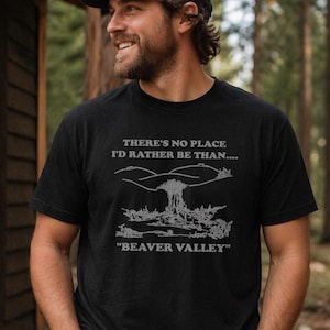May include: Black t-shirt with white text and a landscape graphic. The text reads "THERE'S NO PLACE I'D RATHER BE THAN...." and "BEAVER VALLEY". The person is wearing a black baseball cap. The shirt is made of a soft material.