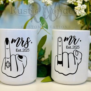 May include: Two white ceramic mugs with black lettering. The mug on the left says "mrs. Est. 2025" with a cartoon hand with a ring on the middle finger. The mug on the right says "mr. Est. 2025" with a cartoon hand with a ring on the middle finger.