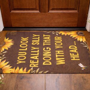 May include: Brown doormat with yellow text that reads "YOU LOOK REALLY SILLY DOING THAT WITH YOUR HEAD...". The mat is decorated with sunflowers and a bee graphic. Placed in front of a wooden door.