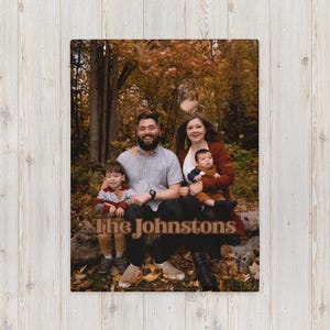 May include: A family photo blanket with a picture of a family of four in a forest setting. The blanket has the text "The Johnstons". The background shows autumn leaves. The blanket is a cozy home decor item.