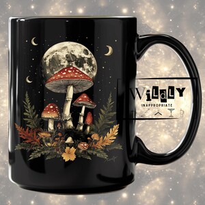 Może przedstawiać: Czarny ceramiczny kubek z fantastyczną ilustracją grzybów pod pełnią księżyca i gwiazdami. Projekt zawiera grzyby z czerwonymi kapeluszami z białymi kropkami, paprocie i jesienne liście. Kubek ma duży uchwyt i napis "WiLaLy INAPPROPRIATE".