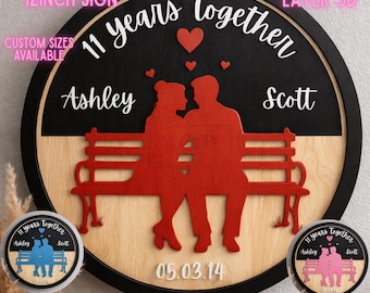 May include: A round, multi-layered wooden sign with a black and natural wood finish. The sign features a red silhouette of a couple on a bench, the text "11 years Together", and the names "Ashley" and "Scott". The sign is 12 inches in diameter.