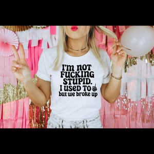Könnte beinhalten: Weißes T-Shirt mit dem Text "I'M NOT FUCKING STUPID. I USED TO but we broke up" in schwarzer, fetter Schrift. Das Shirt hat hochgekrempelte Ärmel und wird von einer Person getragen. Der Hintergrund umfasst rosa und goldene Dekorationen und einen weißen Ballon.
