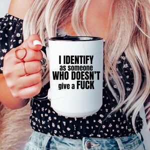 Op de afbeelding: Witte keramische mok met een zwarte binnenkant en handvat. De mok heeft de tekst "I IDENTIFY as someone WHO DOESN'T give a FUCK" in vette zwarte letters. De mok wordt door een persoon vastgehouden.