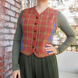 May include: A woman wearing a red and blue checked waistcoat over a long-sleeved green shirt and a dark green pleated skirt. She is standing in front of a brick wall and a window.