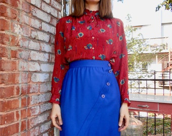 May include: A woman wearing a red and white patterned blouse with a blue skirt and buttons. The skirt has a diagonal seam and is knee-length.