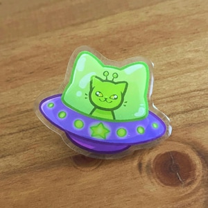 May include: A clear acrylic pin featuring a cartoon cat with antennae inside a bright green cockpit. The cat is piloting a purple UFO saucer decorated with a green star and small green circular lights along the outer edge of the craft.