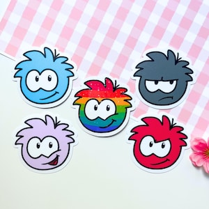May include: A collection of five cartoon stickers featuring round, fluffy characters with expressive eyes and smiles. The stickers are in various colors: blue, rainbow, gray, purple, and red. The background is a pink and white checkered pattern.