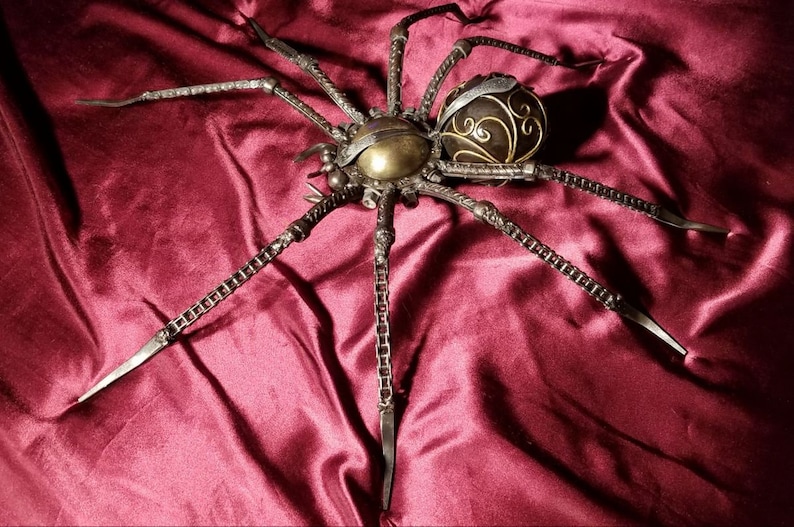 May include: A large, metal spider sculpture with a detailed, ornate body. The spider is made of chains and metal pieces, and has a dark brown and gold finish.