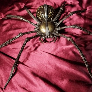 May include: A large, black metal spider sculpture with a gold and black patterned body. The spider is posed with its legs extended, as if it is crawling. The sculpture is set against a red velvet background.