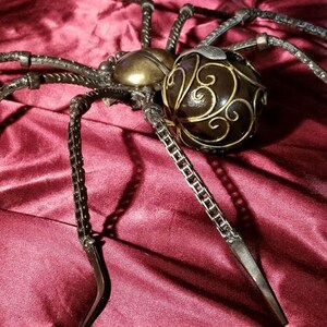 May include: A large, metal spider sculpture with a detailed, ornate, brown and gold body. The spider is made of chains and metal rods and is posed on a red velvet surface.