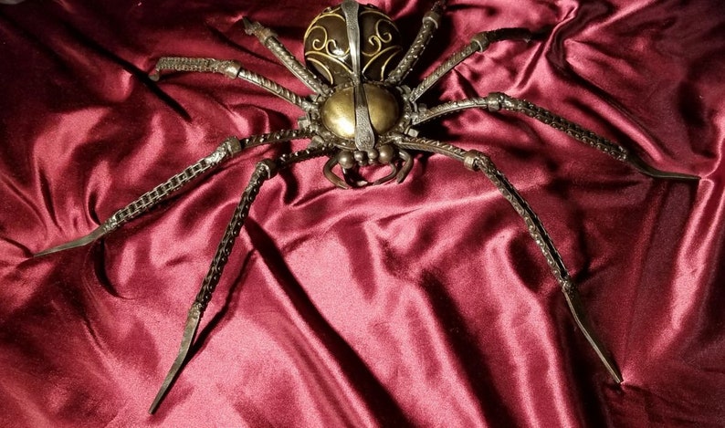 May include: A large, metal spider sculpture with a detailed, ornate design. The spider is made of black metal and has a gold-colored body. The spider is sitting on a red velvet surface.
