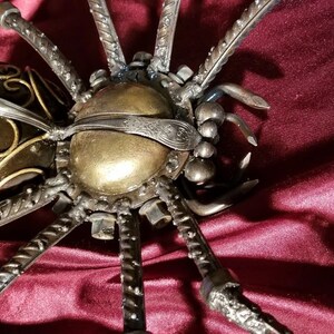 May include: A large, metal spider sculpture with a gold and black body and eight legs. The spider is made of various metal pieces, including screws, nuts, and bolts. The spider is sitting on a red velvet surface.
