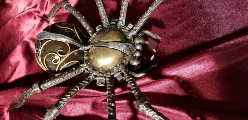 May include: A large, metal spider sculpture with a gold and black body and eight legs. The spider is made of various metal pieces, including screws, nuts, and bolts. The spider is sitting on a red velvet surface.
