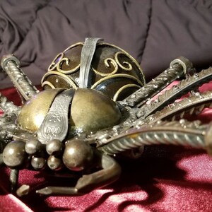 May include: A large, metal spider sculpture with a detailed, ornate design. The spider is made of twisted metal rods and has a black and gold finish.
