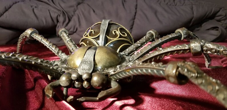 May include: A large, metal spider sculpture with a detailed, ornate design. The spider is made of twisted metal rods and has a black and gold finish.